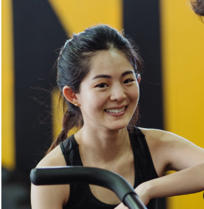 Eine lächelnde Frau in Sportkleidung sitzt in einem Fitnessstudio vor einem gelben und schwarzen Hintergrund.