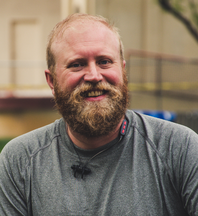 Ein lächelnder Mann mit Bart und grauem Shirt steht draußen, ein Headset um den Hals in einem unscharfen Hintergrund.