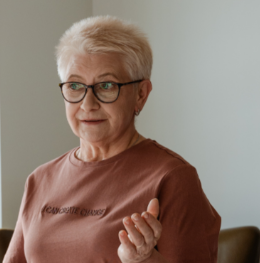 Ältere Frau mit kurzen weißen Haaren und Brille trägt ein braunes T-Shirt und gestikuliert mit der rechten Hand.