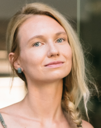 Eine Frau mit blondem Haar und einem geflochtenen Zopf schaut in die Kamera, vor einem unscharfen Hintergrund.