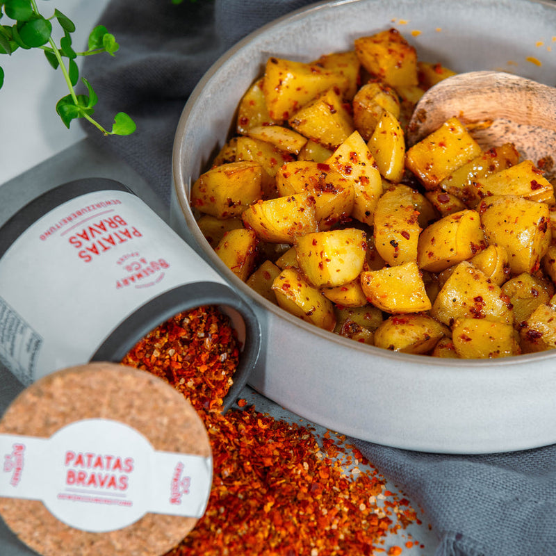Gewürzte Kartoffelstücke in einer Schüssel, daneben ein Glas mit gewürztem Pulver, das Patatas Bravas Gewürz enthält.