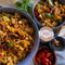 Schüssel mit Farfalle-Pasta, Tomaten, Oliven und Kräutern, daneben Gewürzdosen und frische Tomaten auf einem Tisch.