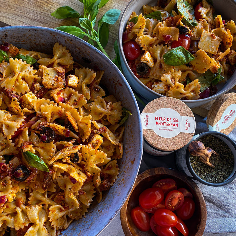 Schüssel mit Farfalle-Pasta, Tomaten, Oliven und Kräutern, daneben Gewürzdosen und frische Tomaten auf einem Tisch.