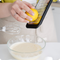 Eine Person reibt eine Zitrone über eine Glasschüssel mit Teig, daneben ein Handmixer und ein geknacktes Ei.