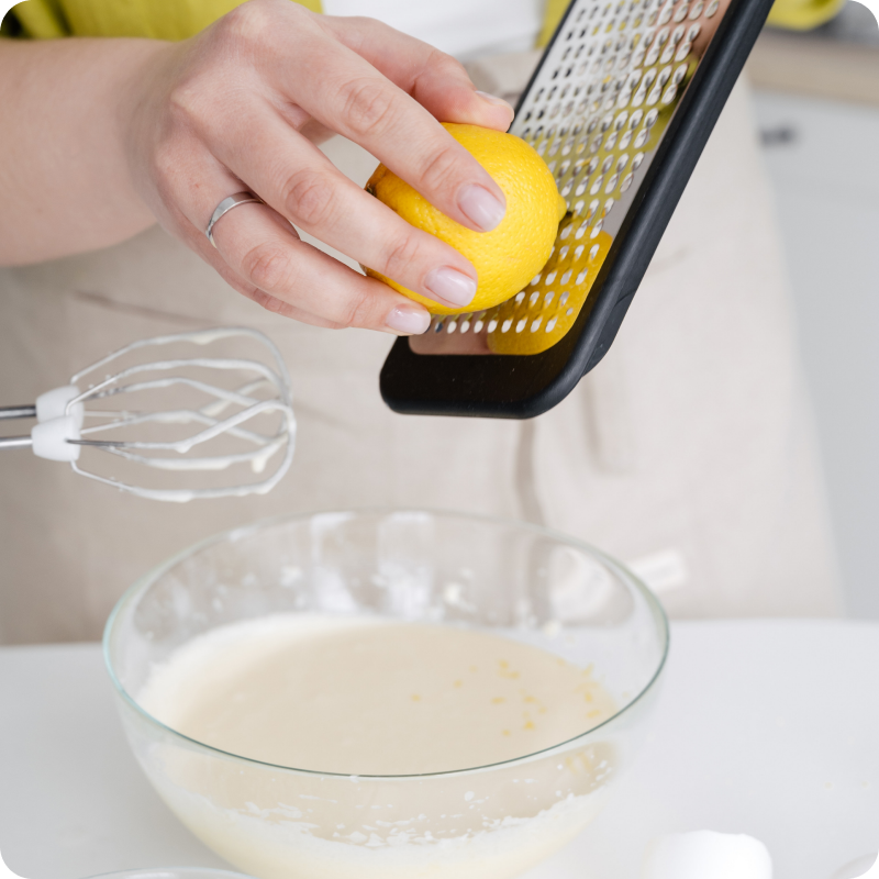 Eine Person reibt eine Zitrone über eine Glasschüssel mit Teig, daneben ein Handmixer und ein geknacktes Ei.