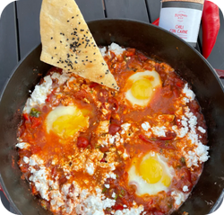 Pfanne mit Shakshuka, drei pochierte Eier und ein Brotstück mit schwarzem Sesam, daneben eine Chilipaste-Flasche.