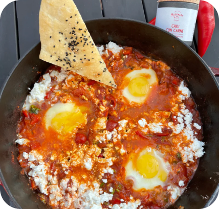 Pfanne mit Shakshuka, drei pochierte Eier und ein Brotstück mit schwarzem Sesam, daneben eine Chilipaste-Flasche.