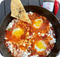 Pfanne mit Shakshuka, drei pochierte Eier und ein Brotstück mit schwarzem Sesam, daneben eine Chilipaste-Flasche.