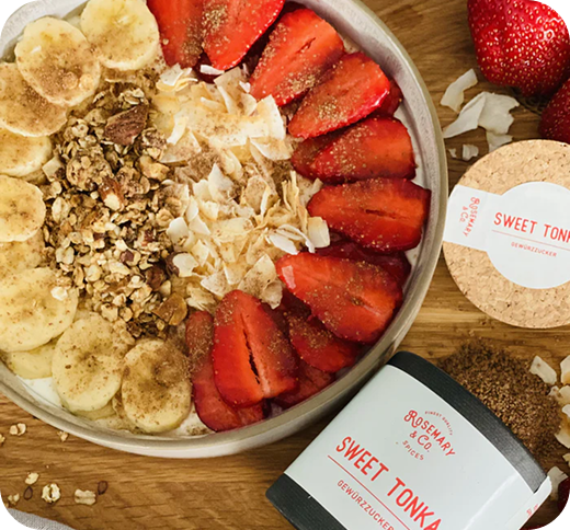 Schale mit Joghurt, Bananen, Erdbeeren, Müsli und Kokosraspeln, umgeben von „Sweet Tonka“ Gewürzzucker auf Holzuntergrund.