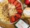 Schale mit Joghurt, Bananen, Erdbeeren, Müsli und Kokosraspeln, umgeben von „Sweet Tonka“ Gewürzzucker auf Holzuntergrund.