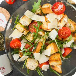 Ein Teller Panzanella-Salat mit Rucola, Tomaten, Croûtons und Parmesanscheiben, daneben Olivenöl und Gewürze.