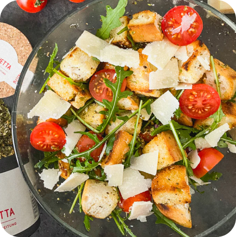Ein Teller Panzanella-Salat mit Rucola, Tomaten, Croûtons und Parmesanscheiben, daneben Olivenöl und Gewürze.