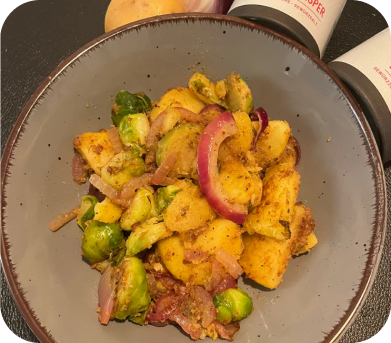 Graue Schale mit Kartoffeln, Rosenkohl, roten Zwiebeln und knusprigem Speck auf schwarzem Untergrund.
