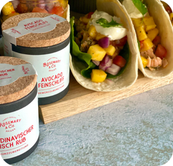 Verschiedene Gewürzdosen neben Tacos, gefüllt mit Mango und Gemüse, auf einem Holztisch.