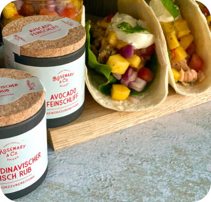Verschiedene Gewürzdosen neben Tacos, gefüllt mit Mango und Gemüse, auf einem Holztisch.